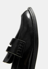 Črna lakasta usnjena čevlja z okroglim prstom, gladko teksturo in značilnim trakom z detajlno šivanko čez zgornji del.