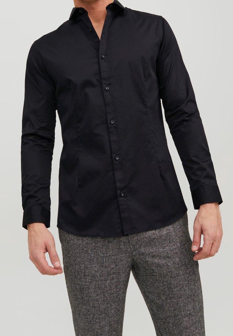 Homme portant une chemise noire ajustée à manches longues et des pantalons gris texturés sur un fond uni.