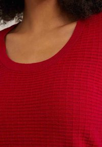 Haut en maille texturée rouge avec un large col et des bords côtelés, présentant un motif de grille subtil sur le tissu.