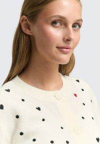 Sweter zapinany na guziki w kolorze off-white z małymi czarnymi sercami i jednym czerwonym sercem. Posiada okrągły dekolt.