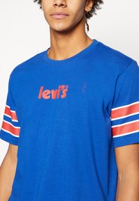 Camiseta de algodón azul con el logo rojo de "levi's", con acentos de rayas blancas y rojas en las mangas. Cuello redondo y mangas cortas.