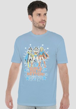 Mann trägt hellblaues T-Shirt mit bunten Cartoon-Superhelden und dem Text "Girls Do It Better" auf der Vorderseite.