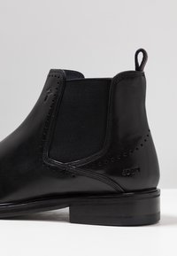 Botín Chelsea de cuero negro con paneles laterales elásticos, perforaciones decorativas y un tacón apilado. Logo de la marca grabado en el tacón.