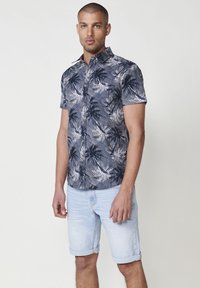 Camisa de botón de manga corta con un patr�ón de hojas de palma en azul marino y blanco, hecha de un tejido ligero. Combinada con pantalones cortos de mezclilla claros.