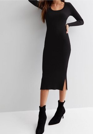 Kvinde bærer en tætsiddende sort midi-kjole med lange ærmer og en sideslids, stylet med sorte ankelstøvler mod en enkel lys baggrund.