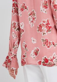 Blusa floreale rosa con un tessuto morbido e leggero. Motivi floreali in rosso, bianco e marrone, con maniche a campana e dettagli con bottoni.