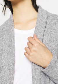 Jasnoszary kardigan o miękkiej fakturze, z otwartym przodem i szerokim kołnierzem. Modelka ma na sobie białą bluzkę pod spodem, trzymając kardigan jedną ręką.