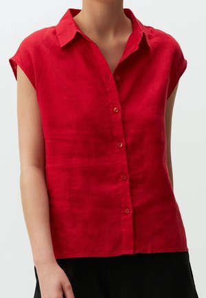 Femme portant une chemise rouge sans manches boutonnée avec col, debout devant un fond clair uni.
