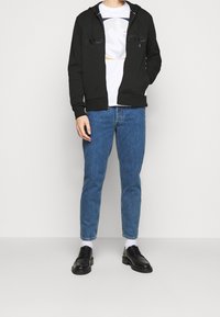 Felpa con cappuccio nera con cerniera, t-shirt bianca con grafica sotto, jeans di denim blu e scarpe nere. La felpa ha coulisse e polsini a coste.