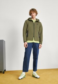 Felpa con cappuccio verde oliva con zip, dettagli neri e tasche, indossata sopra una maglia verde chiaro. Jeans blu e scarpe da ginnastica bianche con dettagli verdi.