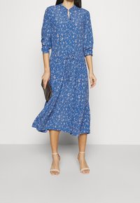 Blå blommig midi-klänning med nyckelhålsringning, långa ärmar, indragen midja och svängande nederdel, i kombination med sandaler med genomskinliga remmar och en svart kuvertväska.