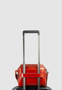 Bolsa de viaje de cuero rojo con múltiples compartimentos con cremalleras, que cuenta con un asa resistente y una correa ajustable, colocada sobre una maleta negra.