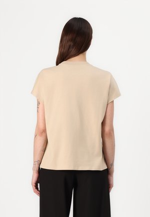 Person med langt brunt hår iført beige kortærmet skjorte og sorte bukser, set bagfra mod en ensartet hvid baggrund.