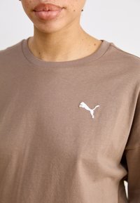 Brūna kokvilnas T-krekls ar īsām piedurknēm, apaļu kaklu un baltu izšūtu Puma logo kreisajā krūtī. Gluda tekstūra.