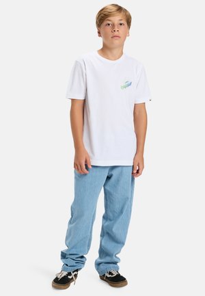 Weißes Baumwoll-T-Shirt mit buntem Grafikdesign auf der Brust, kombiniert mit hellblauen Jeans und schwarzen Freizeitschuhen mit beigen Akzenten.