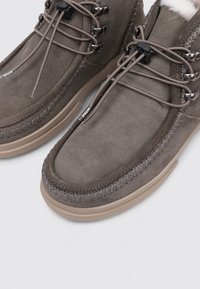 Bottes en suède gris avec des semelles en caoutchouc beige, des coutures texturées contrastantes et des lacets élastiques pour un maintien sécurisé. Doublure douce visible en haut.