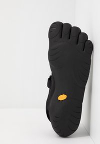 Semelle en caoutchouc noir avec design à cinq orteils, surface texturée et logo Vibram® en orange. Forme flexible avec motifs de traction incurvés.