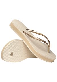Vaaleanbeige flip-flopit, joissa on teksturoitu pohja, metallinhohtoiset kultaiset hihnat ja tasainen pohja, jossa on pyöreä logo.