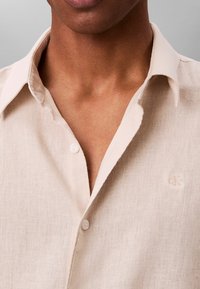 Primer plano de una persona que lleva una camisa de lino beige con el cuello abierto y dos botones abrochados, mostrando parcialmente el pecho y el cuello.