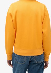 Sudadera amarilla con cuello redondo acanalado y puños, que presenta una textura suave y un corte relajado. Combinada con jeans de mezclilla azul oscuro.