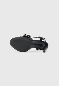 Sort højhælet sandal med ankelrem og dekorative sten, vist fra sålesiden på en ensfarvet lys baggrund.