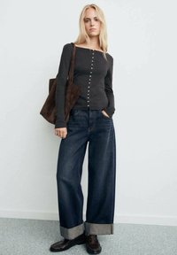 Donkergrijze longsleeve top met knoopdetails, gecombineerd met wijde donkere spijkerbroeken met opgerolde hems en een bruine schoudertas.