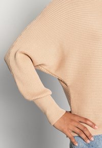 Pullover beige a coste con maniche lunghe e larghe e orlo arrotondato, realizzato in un tessuto morbido con una superficie testurizzata.