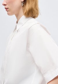 Νεαρό άτομο με ξανθά κοντά μαλλιά, που φοράει ασημένια κρίκους στα αυτιά και λευκό πουκάμισο με κοντά μανίκια και γιακά, κοιτάζοντας προς τα αριστερά μπροστά σε απλό φόντο.