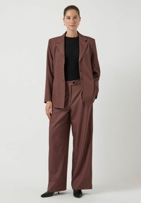 Vrouw in een bruin oversized pak met wijde broekspijpen en een zwart topje, met zwarte puntige hakken, tegen een effen achtergrond.