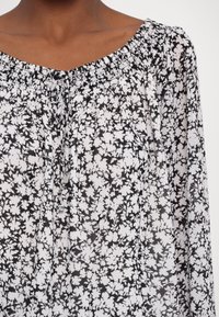 Blouse fleurie en noir et blanc avec un col froncé et un cordon ajustable, portée par une personne avec le cou et le haut de la poitrine visibles.