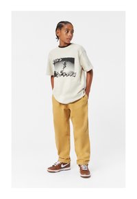 Beez graafiline t-särk musta printiga, koos lõtvade kollaste pükstega. Pruunid tossud valgete aktsentidega täiustavad riietust.