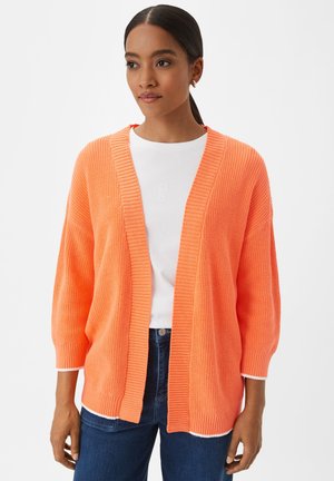 Offenes Strickcardigan in leuchtendem Orange, mit einem Schalkragen, überschnittenen Schultern, gerippten Bündchen und einer entspannten Passform. Über einem weißen Oberteil getragen.
