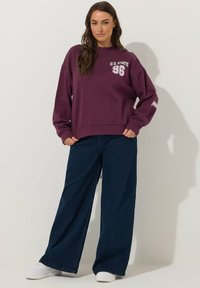 Felpa viola con la stampa "U.S. STATE 96" sul petto. Vestibilità ampia, polsini a coste, abbinata a jeans in denim scuri a gamba larga e sneakers bianche.