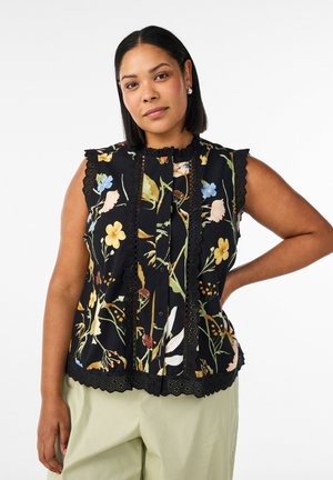 Kvinde med skulderlangt mørkt hår iført en ærmeløs sort bluse med flerfarvet blomsterprint og beige bukser, stående med den ene hånd på hoften.