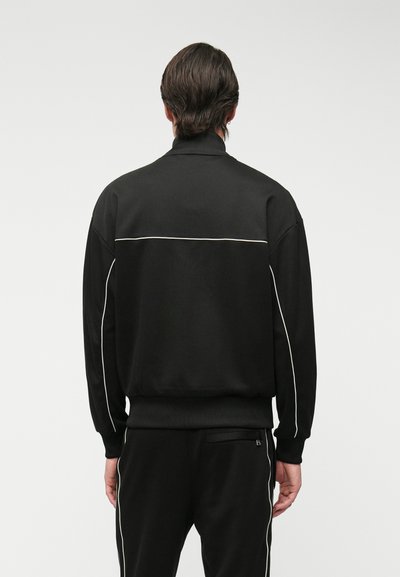Veste de sport noire avec col montant, manches et ourlet côtelés. Des passepoils blancs le long du dos et des côtés ajoutent un contraste au design.