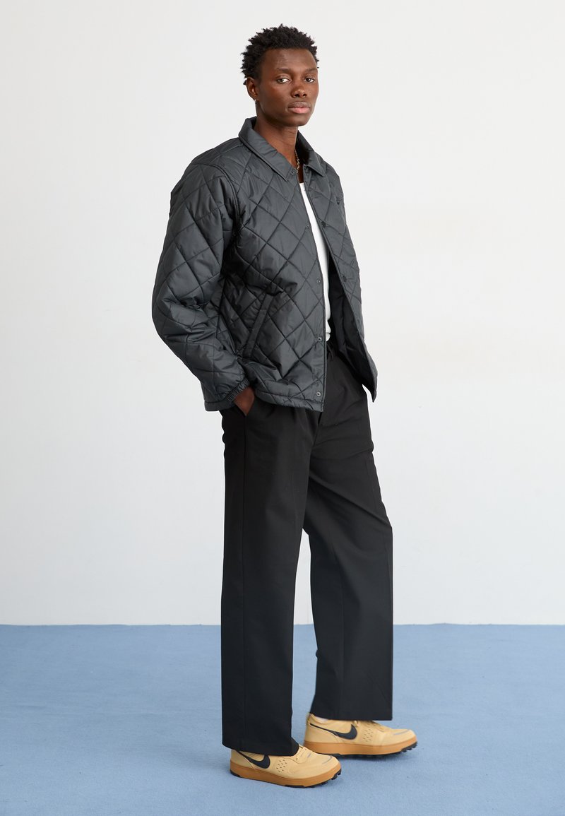Chaqueta acolchada negra con cuello, llevada sobre una camisa blanca. Pantalones negros y zapatillas Nike beige con acentos negros sobre un suelo azul.