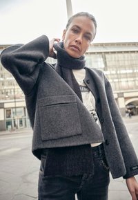 Jachetă gri din lână cu mâneci supradimensionate, buzunar aplicat și închidere cu nasturi, purtată peste un pulover negru tricotat și asortată cu blugi închiși la culoare.