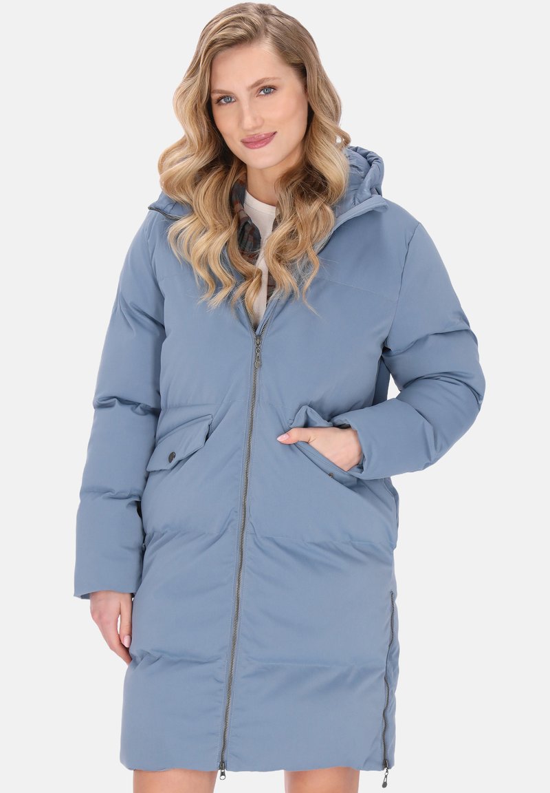 Hosszú, hullámos szőke hajú nő világoskék, kapucnis téli kabátban, elöl cipzárosan, jobb zsebébe dugta a kezét.