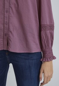 Camicia button-up viola con tessuto testurizzato, maniche a sbuffo e dettagli in pizzo ai polsini. Abbinata a jeans blu scuro.
