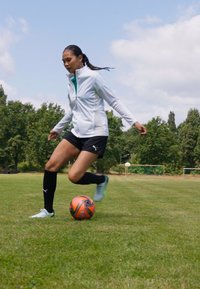 En spiller i en hvit jakke og svarte shorts sparker en oransje og svart fotball på en gressplen, omringet av trær og mål.