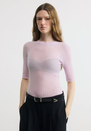 Νεαρή γυναίκα με μακριά καστανά μαλλιά, φοράει ροζ ανοιχτόχρωμο ριμπ τοπ, μαύρο παντελόνι και μαύρη ζώνη, στέκεται μπροστά σε απλό λευκό φόντο.