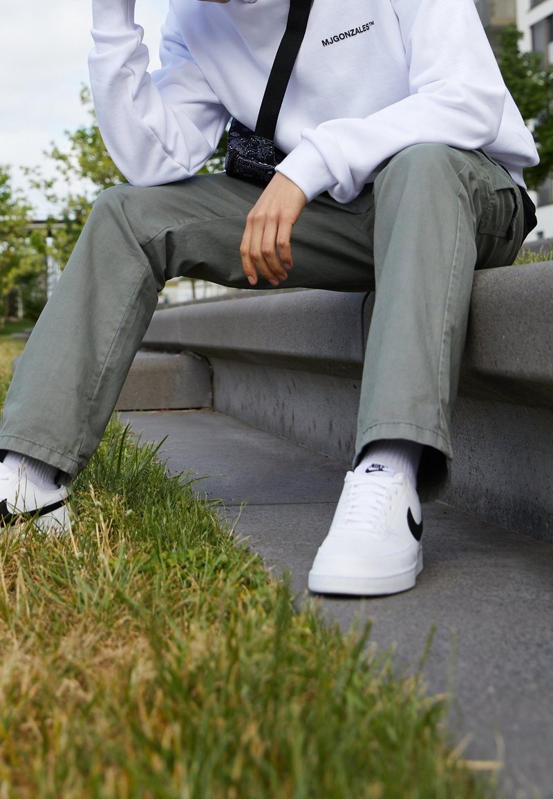 Sweat-shirt blanc, pantalon vert olive et baskets Nike blanches avec des accents noirs. Personne assise sur un rebord en béton près de l'herbe.
