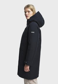 Femme portant un long manteau à capuche noir avec le logo Aigle sur la manche, se tenant de côté sur un fond clair et uni.