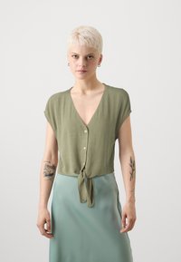 Blusa verde a maniche corte con scollo a V, bottoni frontali e hem annodato. Tessuto liscio e design leggero. Indossata con una gonna verde menta.
