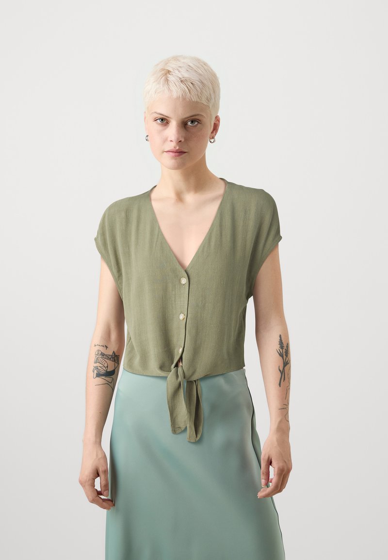 Blusa verde a maniche corte con scollo a V, bottoni frontali e hem annodato. Tessuto liscio e design leggero. Indossata con una gonna verde menta.