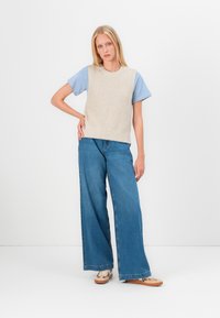 Gilet en tricot beige porté sur une chemise rayée bleu et blanc, associé à un jean en denim bleu à coupe large et des chaussures à enfiler à motifs.