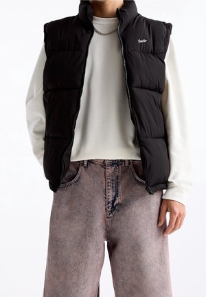 Personne portant un gilet matelassé noir par-dessus une chemise blanche à manches longues et un jean violet délavé, avec un collier chaîne argenté.