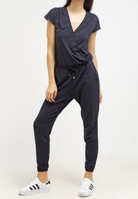 Mono azul marino con un escote en V profundo, mangas cortas, cintura elástica y piernas entalladas. Textura suave; combinado con zapatillas blancas.