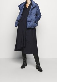 Granatowa kurtka puchowa o błyszczącej powierzchni, z detalami guzików, zestawiona z ciemną sukienką z plisowanym dołem i wysokimi czarnymi botkami.