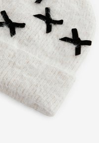 Bonnet tricoté blanc avec une texture duveteuse, comportant des motifs en X en velours noir et un revers replié en bas.